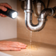 Finding a plumbing leak under a kitchen sink using a flashlight with water dripping from a pipe